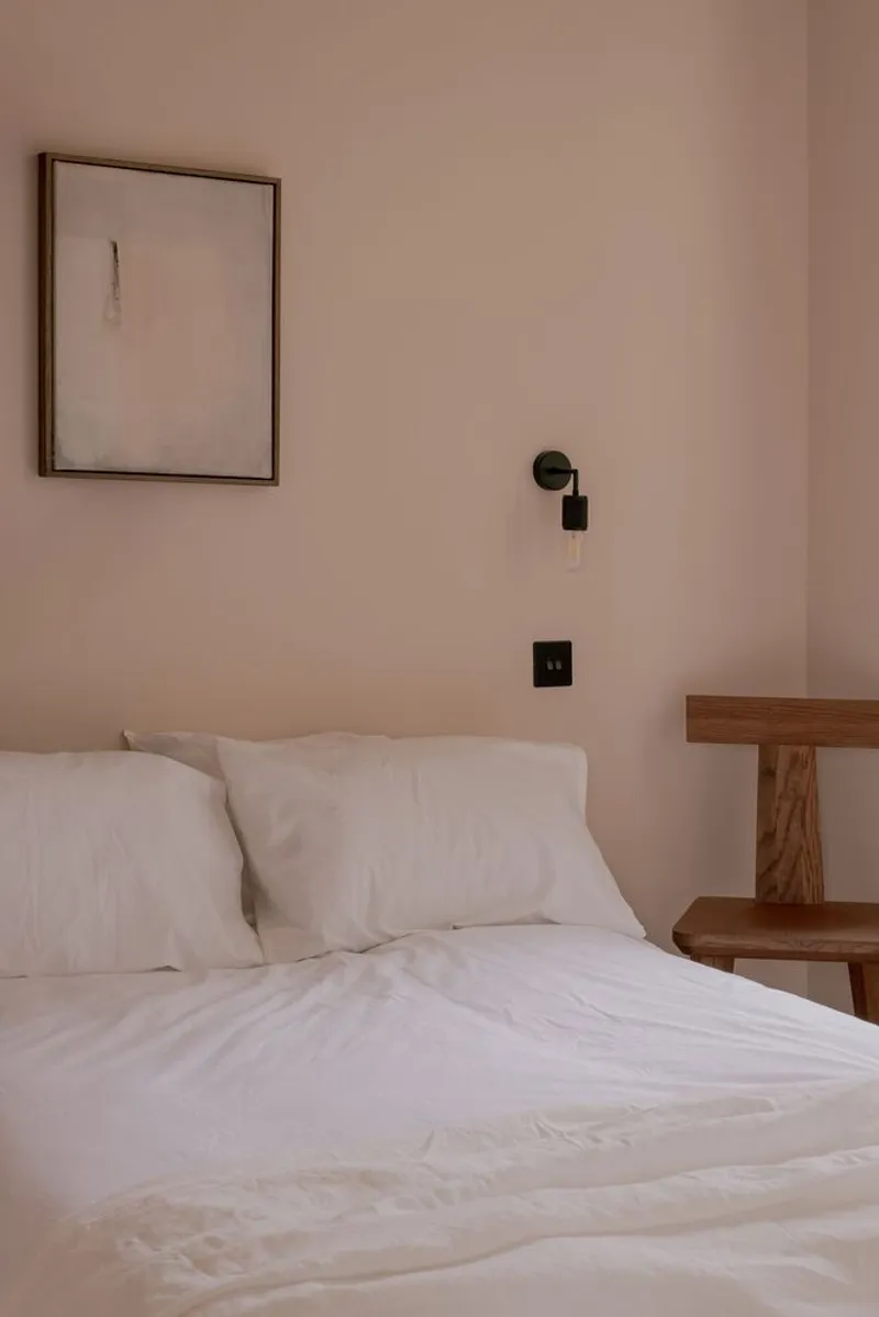 Bedroom with pink plaster walls, white linen bedding, black wall sconce and timber side table