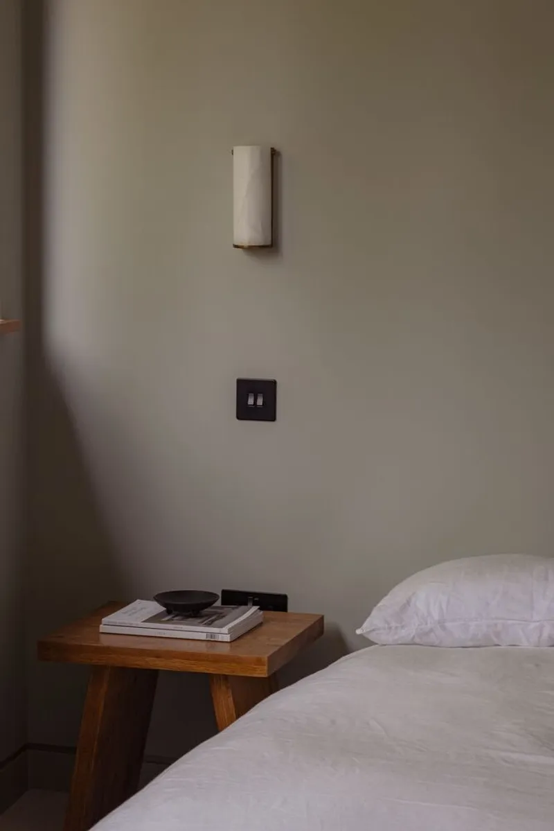 Detail of bedside wall showing black power outlets, cylindrical wall sconce and timber nightstand with books