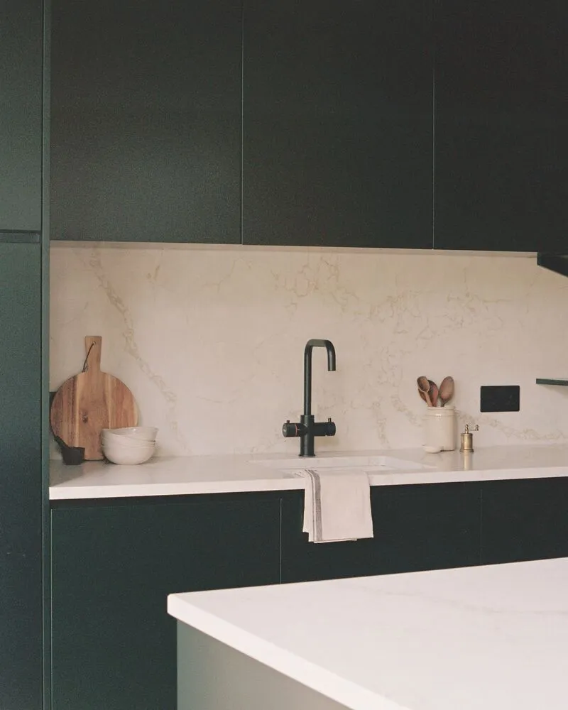 Kitchen counter with dark green cabinets, pale marble backsplash and black tapware with timber cutting boards