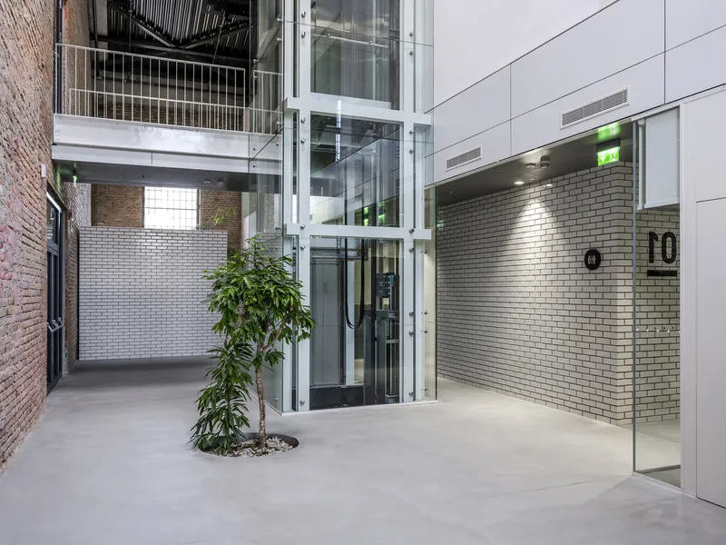 Glass elevator shaft and polished concrete floor with potted tree beside white tile entry wall