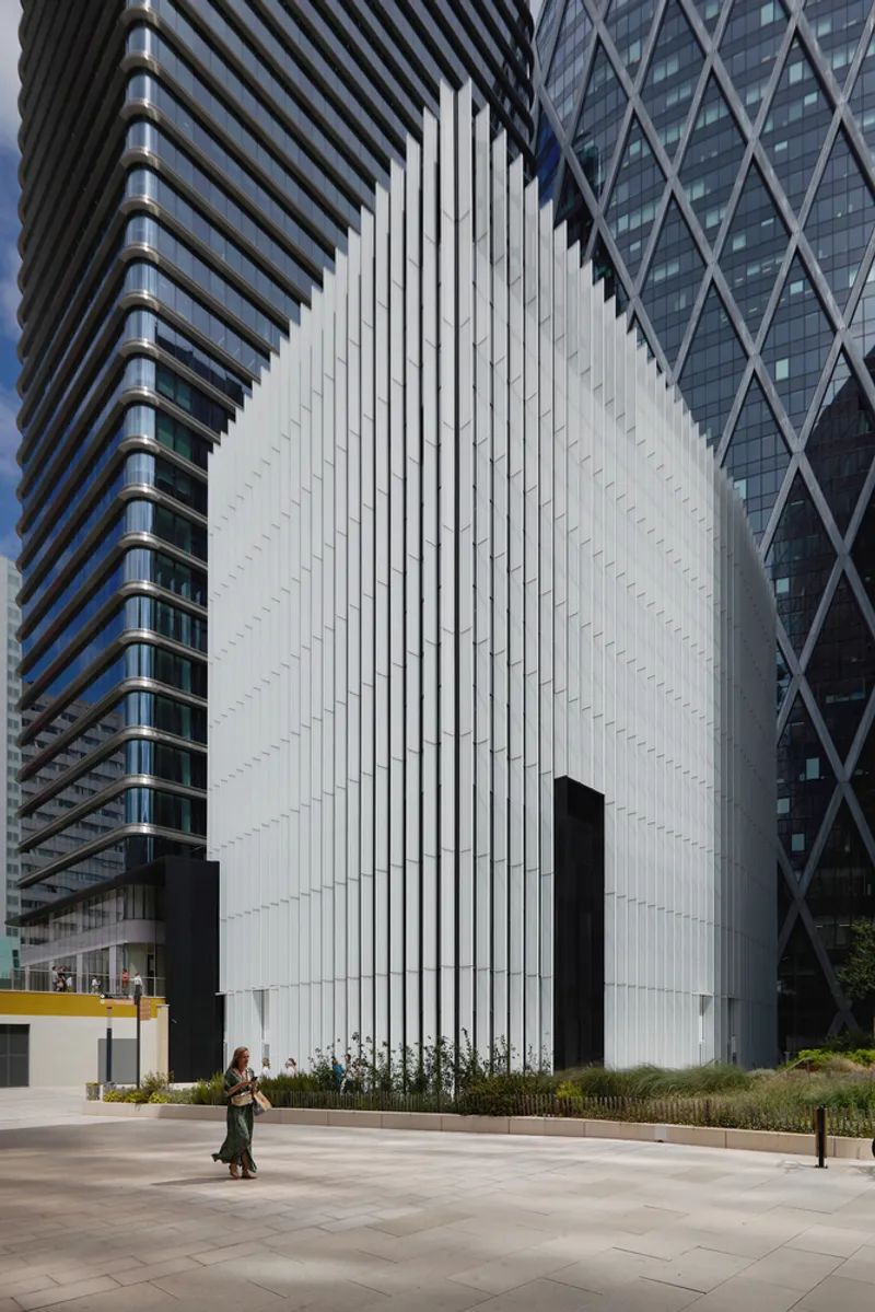 White vertically ribbed facade set between two high-rise towers as a pedestrian passes on the plaza