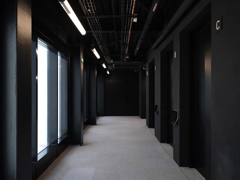Blackened corridor with exposed ceiling services and strip lighting running past window bays and door entries
