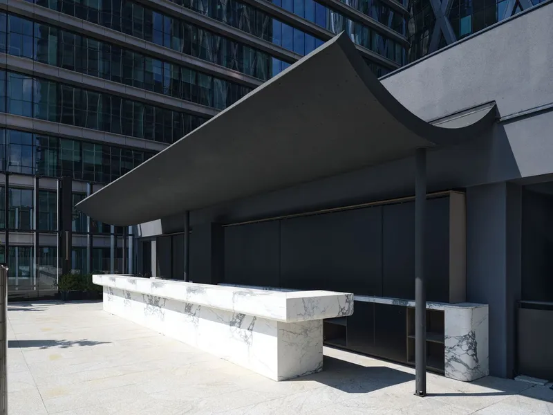 White marble bench beneath a dark canopy supported by cylindrical columns in bright sunlight