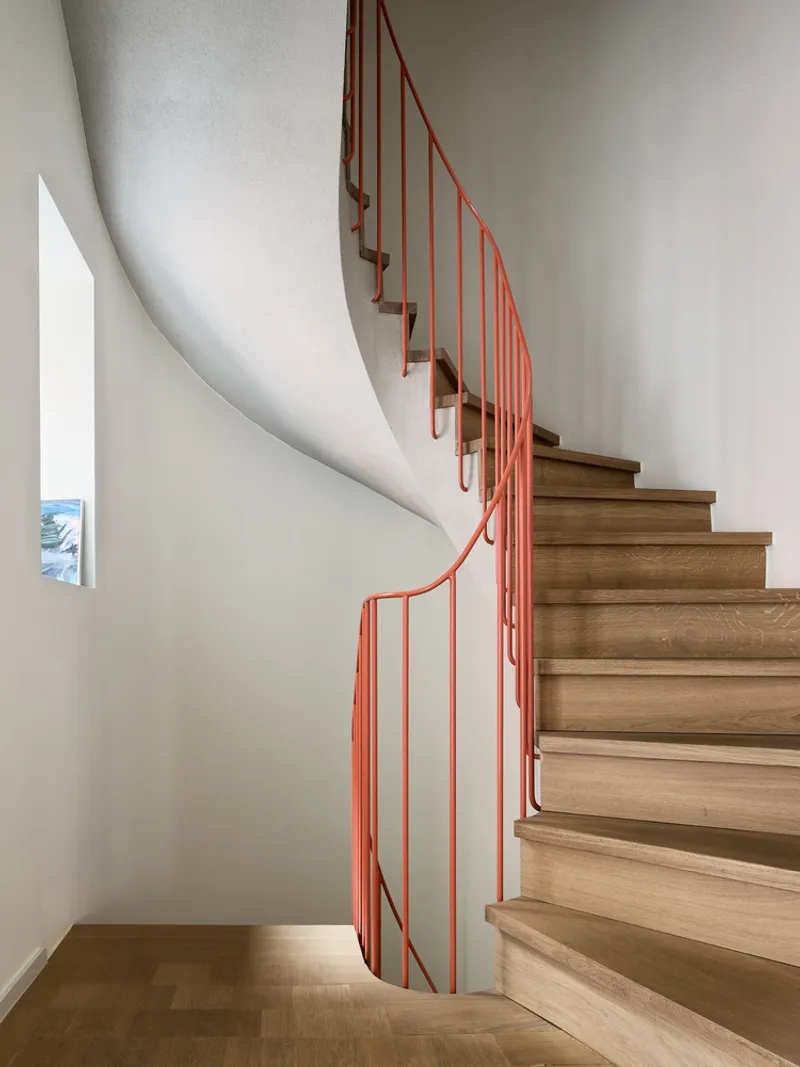 Curving timber staircase with coral-colored metal railing against white walls and wood flooring