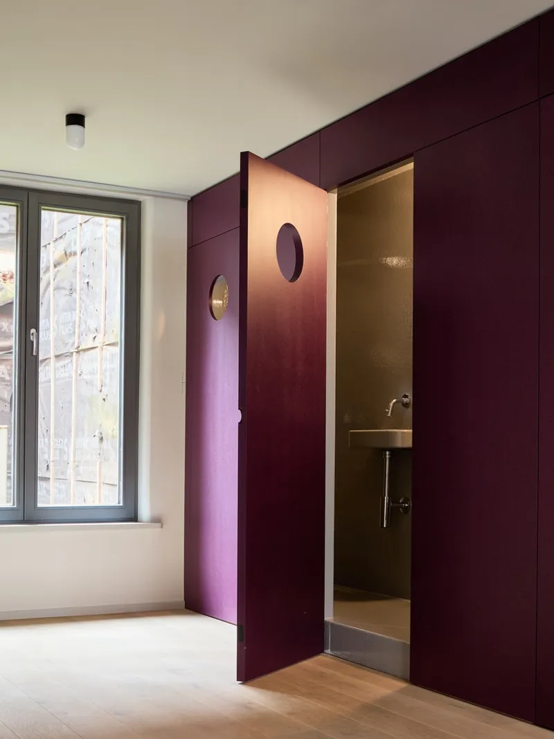 Deep burgundy paneled wall with pivoting door revealing a compact bathroom with brass fixtures