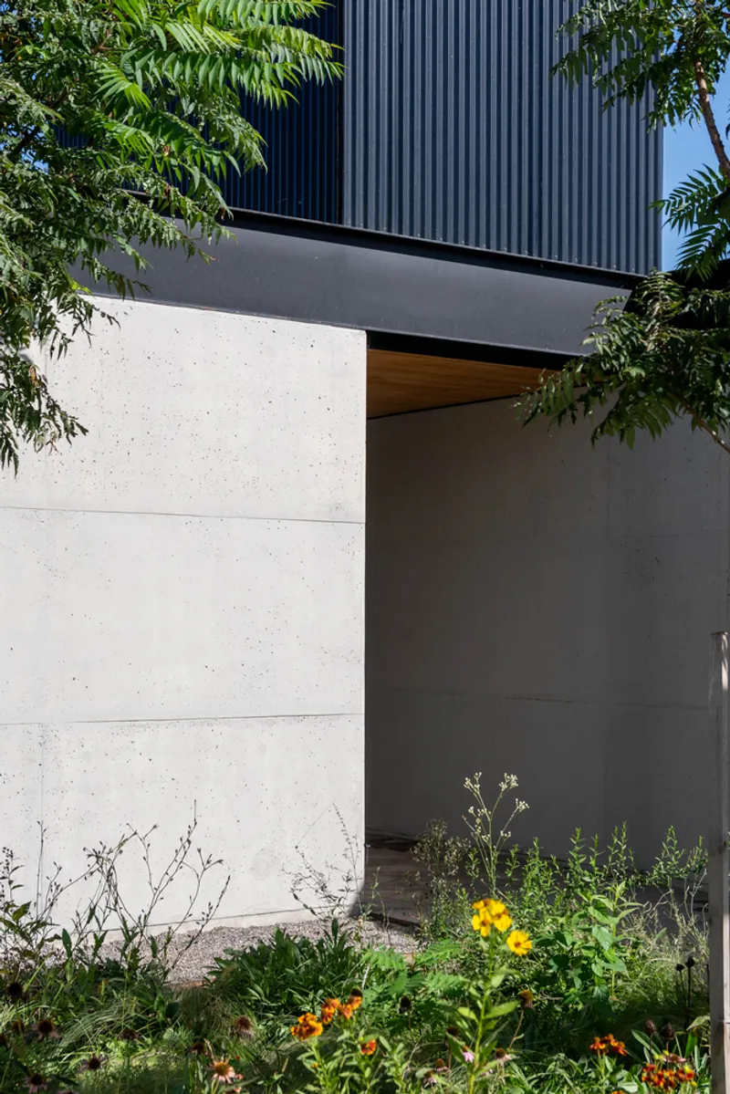Entrance niche in concrete facade with corrugated upper volume and flowering garden bed