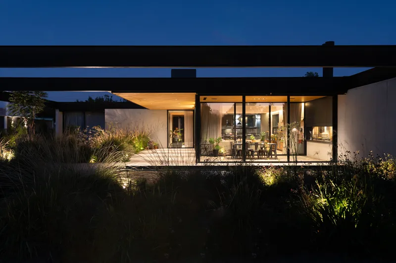 Courtyard view at dusk showing glazed living spaces under steel frame and planted beds with uplighting