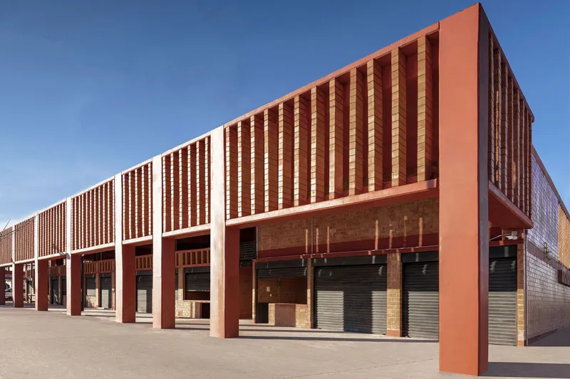 Linear facade with vertical brick banding and steel columns fronting closed roller shutters in bright sun