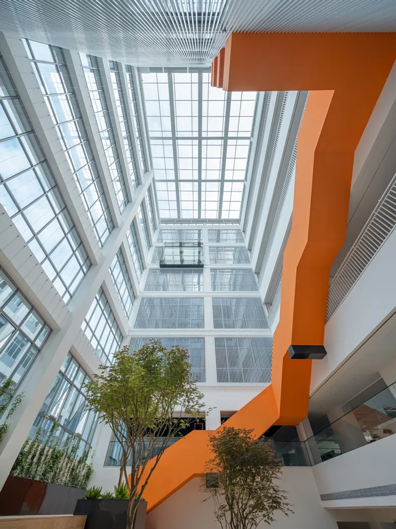 Upward view of the atrium showing orange zigzag stairs beneath a glazed skylight and potted trees below