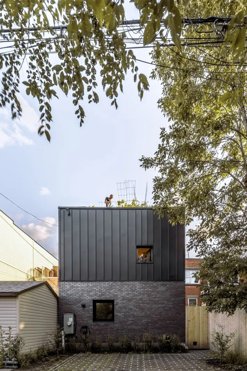 Street view of the two-story volume with dark metal cladding over brick base beneath overhanging birch branches