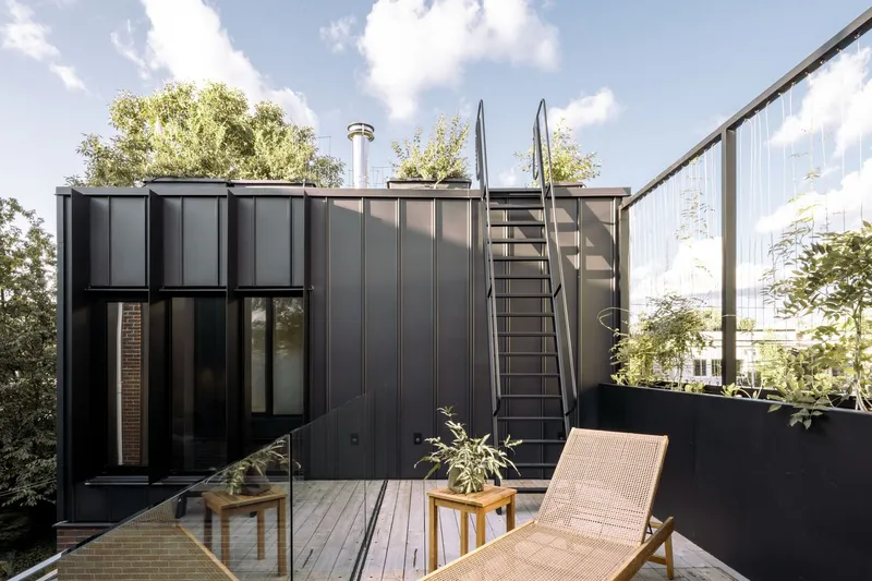 Rooftop deck with metal stair ladder and rattan lounger surrounded by planted screens and neighboring houses