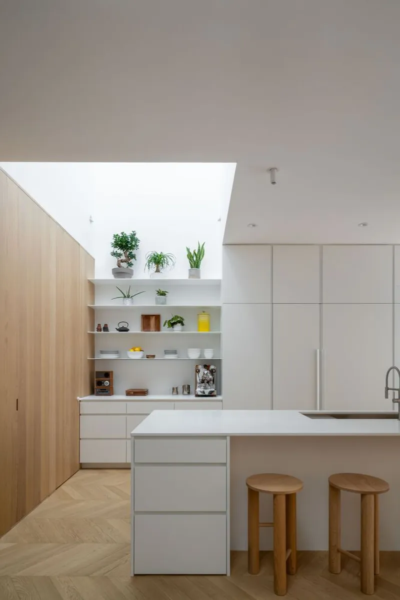 Kitchen island with white countertop and timber stools beneath a rectangular skylight with potted plants