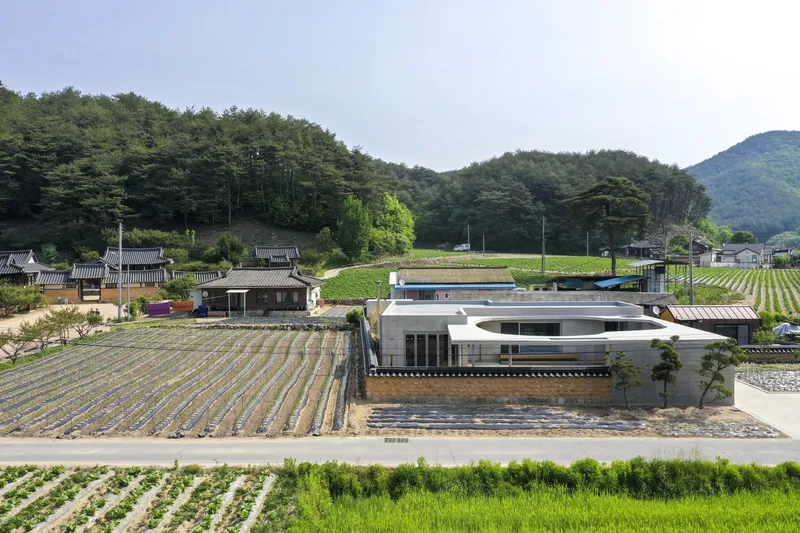 Aerial view of the white-roofed residence nestled among traditional village houses and agricultural fields beneath forested hills
