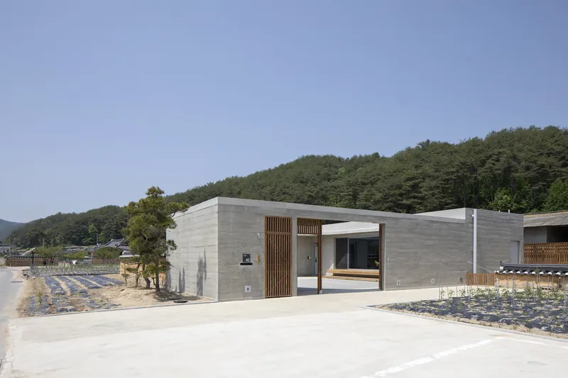 Board-formed concrete entrance volume with vertical timber slat cladding facing the adjacent planted field