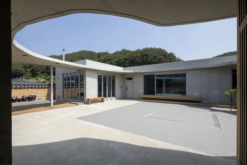 View through curved canopy opening toward central courtyard with timber benches and concrete volumes