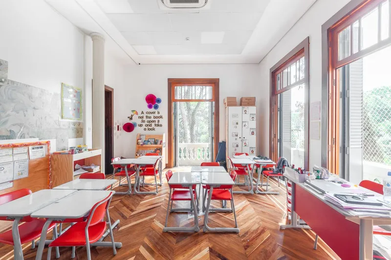 Classroom interior with herringbone wood flooring and red chairs arranged around white work tables beneath tall windows