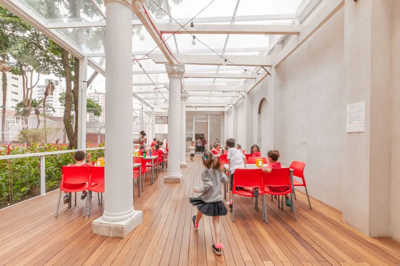 Covered outdoor terrace with timber decking and white columns as children in motion blur pass by red seating