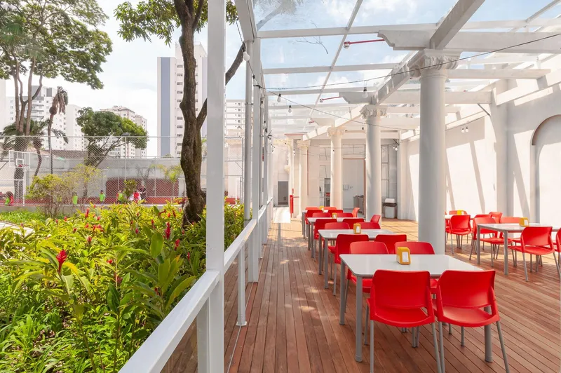 Open-air timber deck dining area with red furniture alongside a planted garden bed under a white pergola structure