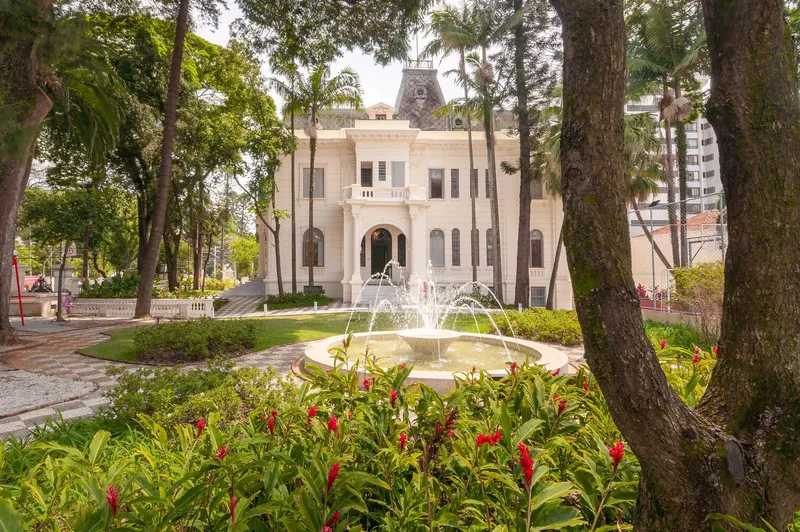 Garden view of a two-story symmetrical facade with arched entry through mature trees and a central fountain