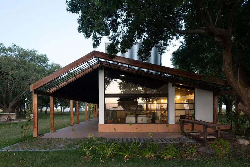 Timber-framed pavilion with extended roof and glass facade beneath mature trees at dusk