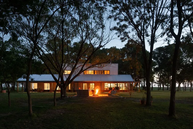 Two-story volume with illuminated windows nestled among trees with a bonfire in the foreground