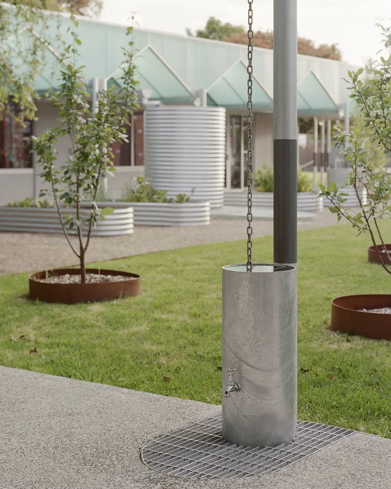 Concrete rainwater chain column with tap and steel grate in a lawn with planted beds and corrugated metal beyond