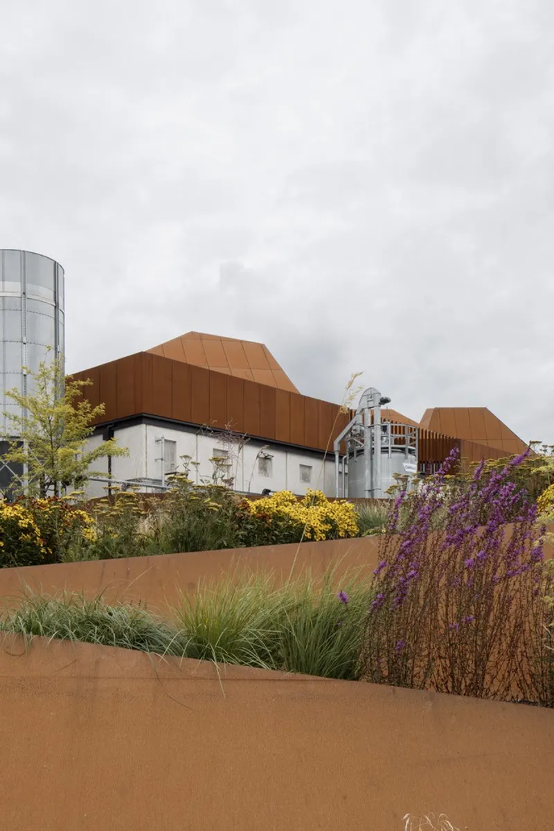 View across planted beds with purple flowering grasses toward the faceted weathered steel roof canopy