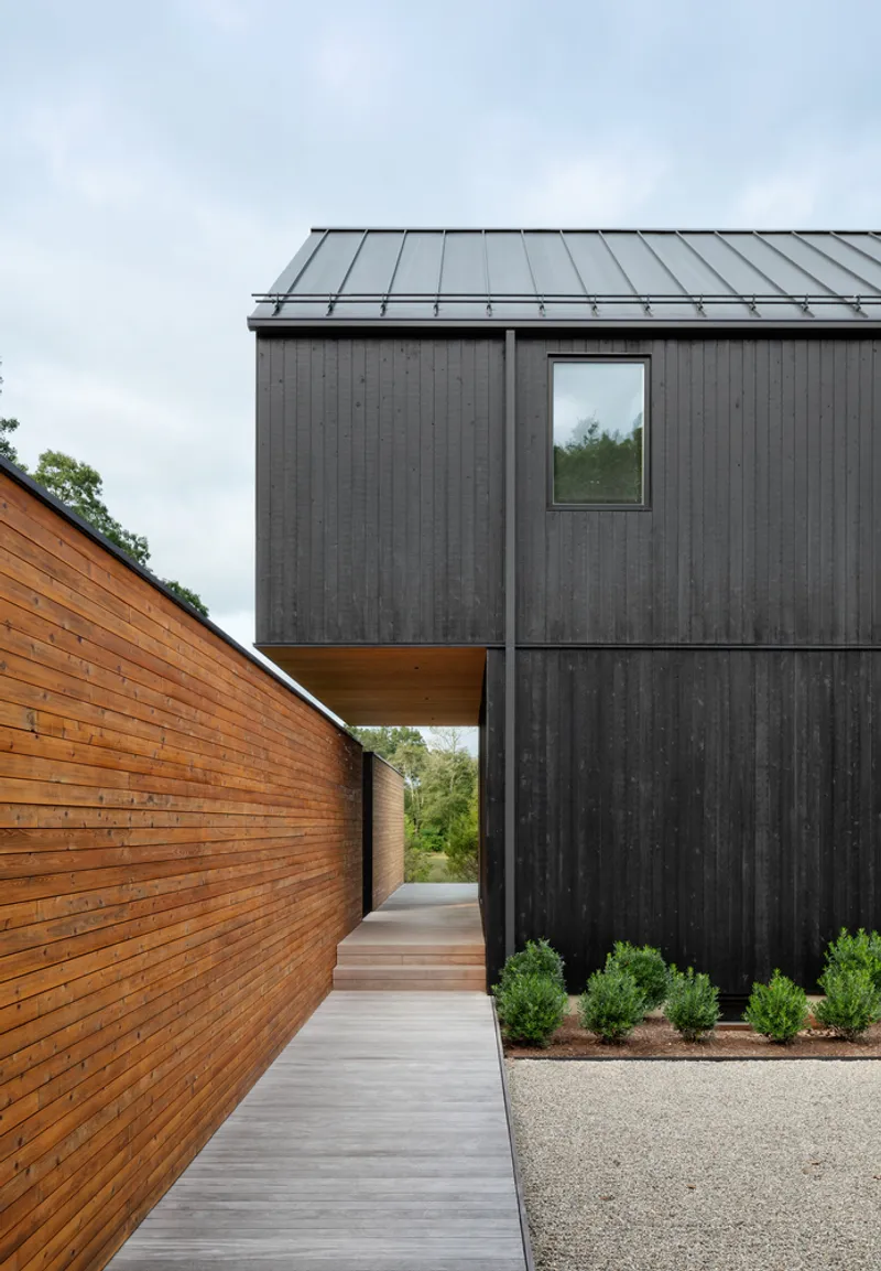 Covered walkway between charred black siding and natural cedar wall under a cantilevered upper volume