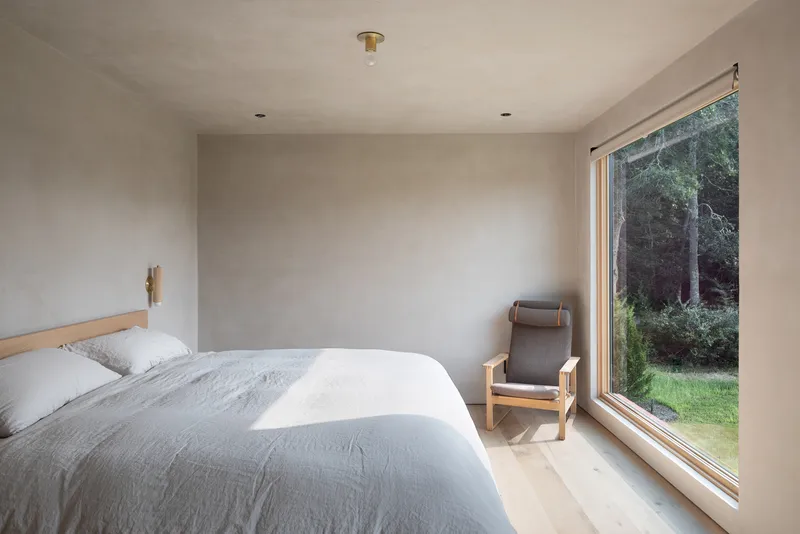 Bedroom with floor-to-ceiling timber-framed window overlooking a forested landscape in soft afternoon light