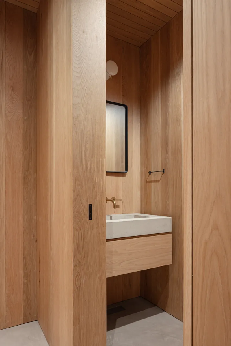 View through vertical oak paneling into a compact bathroom with integrated sink and black mirror