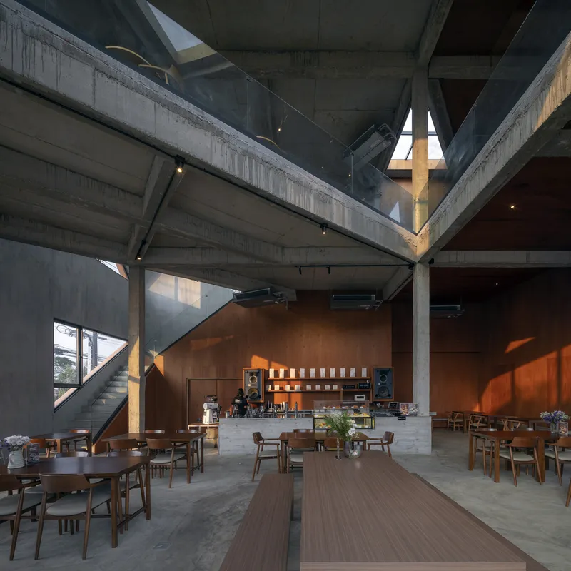 Double-height cafe space with exposed concrete frame, timber-clad walls and triangular skylights casting angular shadows
