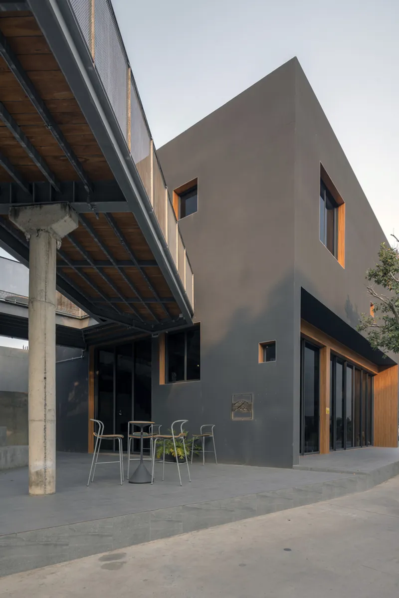 Grey rendered volume with timber-framed window openings beneath an elevated steel and timber walkway at dusk