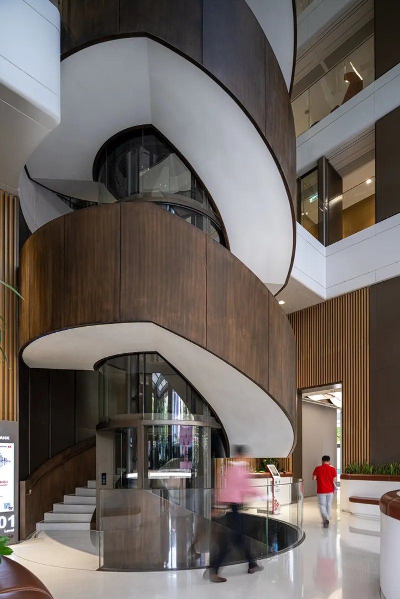Spiraling white staircase with curved soffits wrapping around a cylindrical timber volume in a multi-story atrium