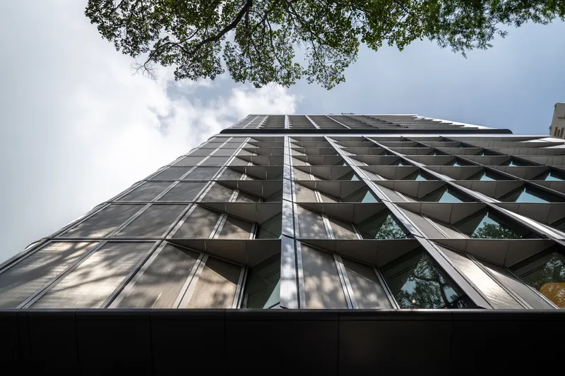 Upward view of the triangular faceted facade with tree branches overhead against cloudy sky