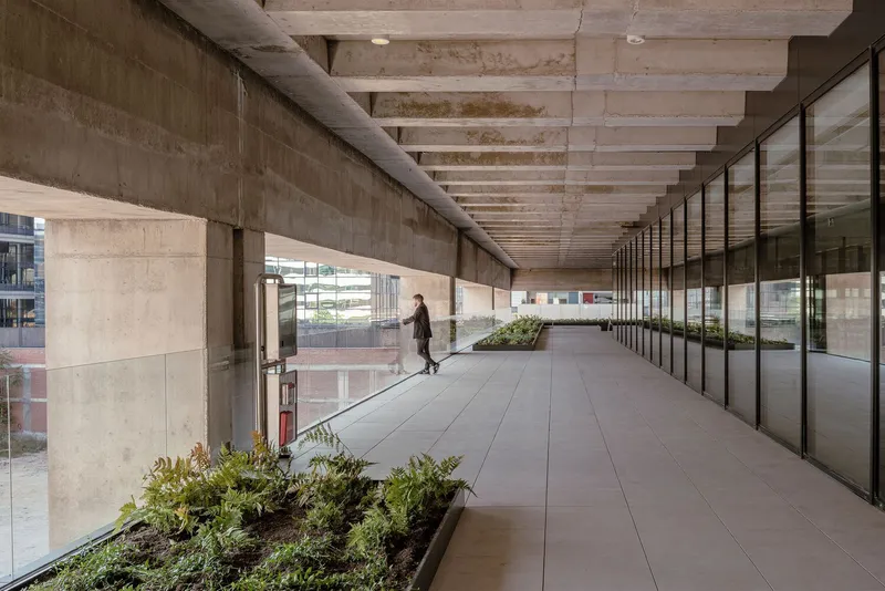 Covered walkway with ribbed concrete soffit, planted beds, and a person walking in shadow