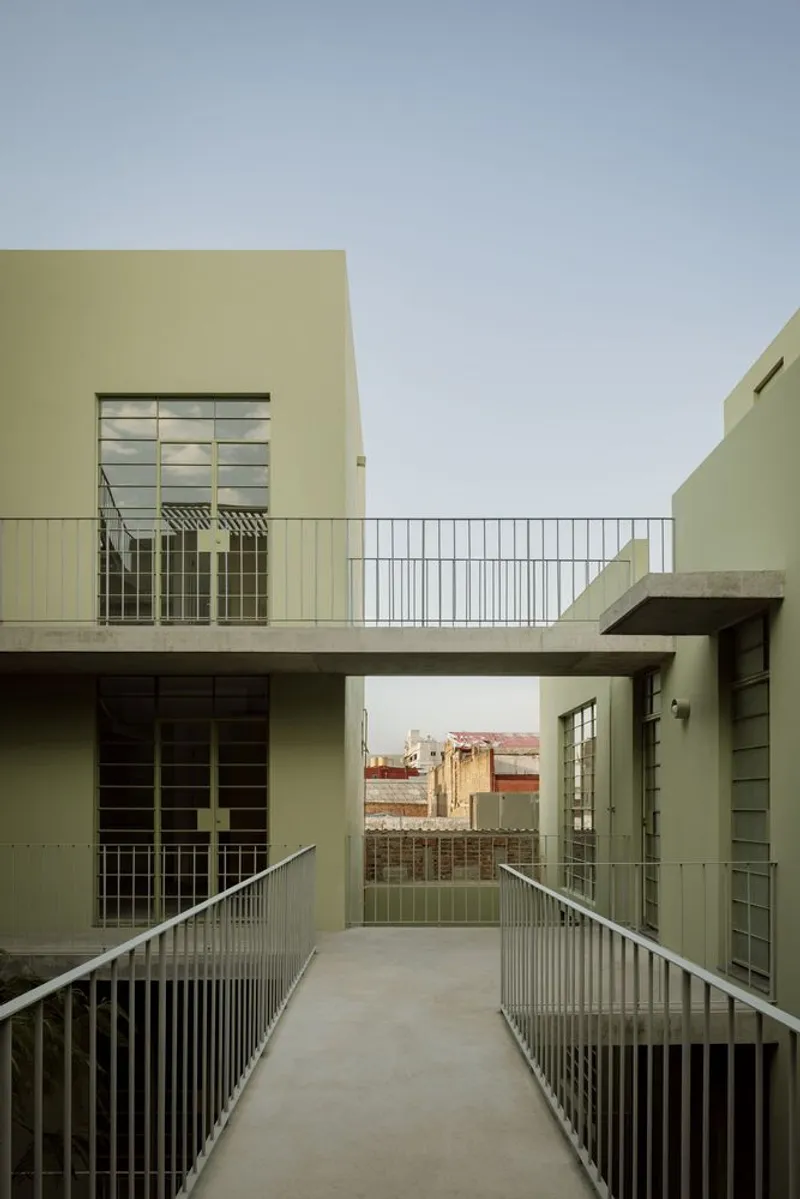 Concrete bridge connecting two pale yellow volumes across an open-air courtyard with distant rooftops visible
