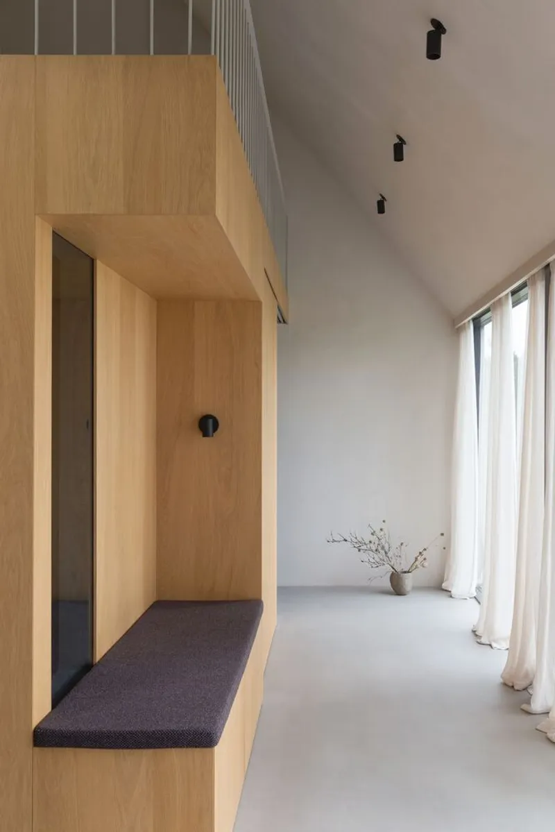 Plywood-clad entry nook with integrated bench beneath a pitched white ceiling and sheer curtains