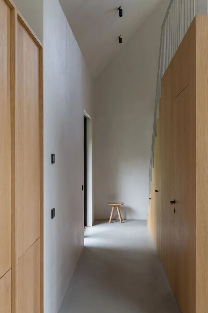 Narrow corridor with oak cabinetry and white walls leading to a wooden stool bathed in light