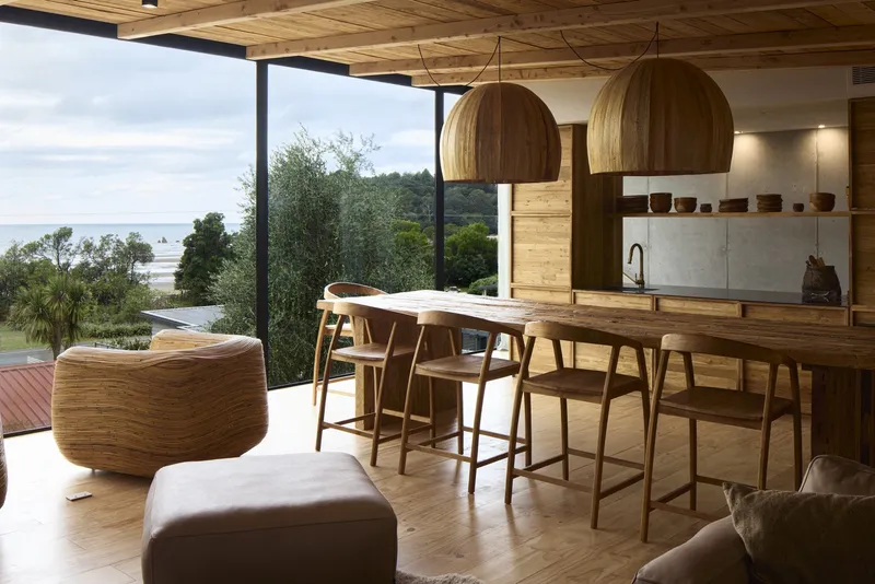 Open plan dining area with timber slatted ceiling and floor-to-ceiling glazing framing coastal vegetation
