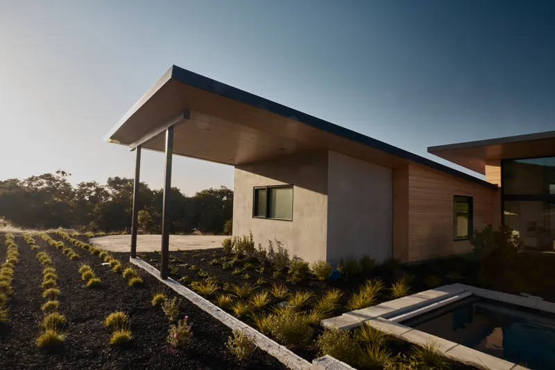 Exterior view of cantilevered roof over stucco and timber volumes with planted beds at dusk