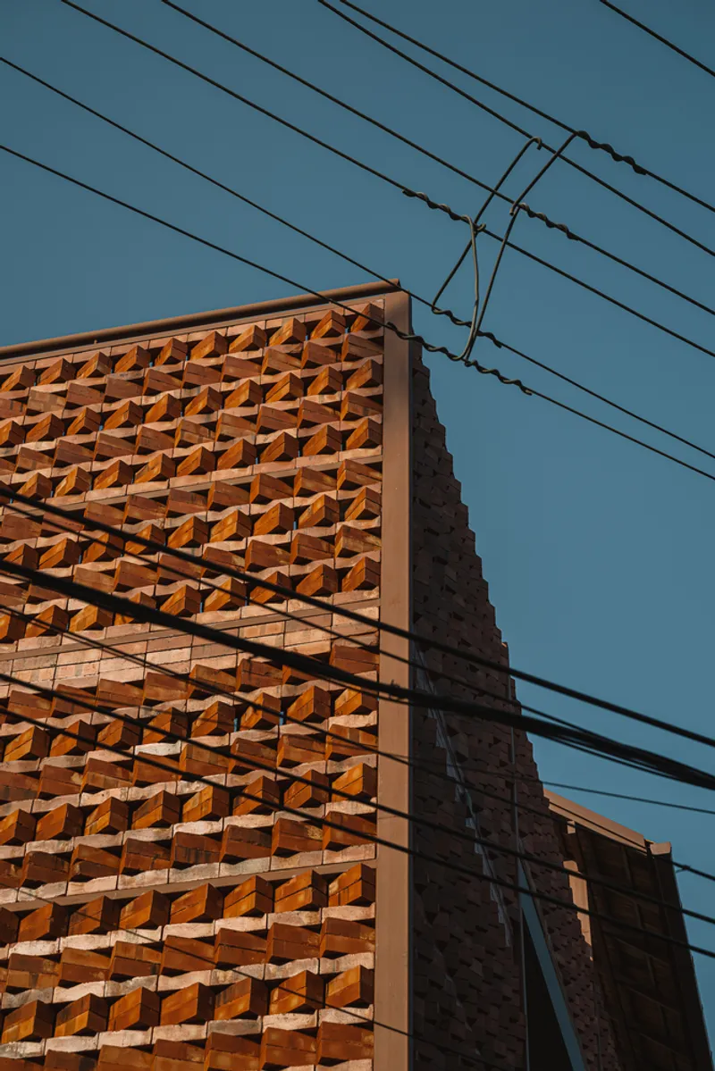 Terracotta brick facade with folded geometric pattern intersected by overhead power lines against blue sky