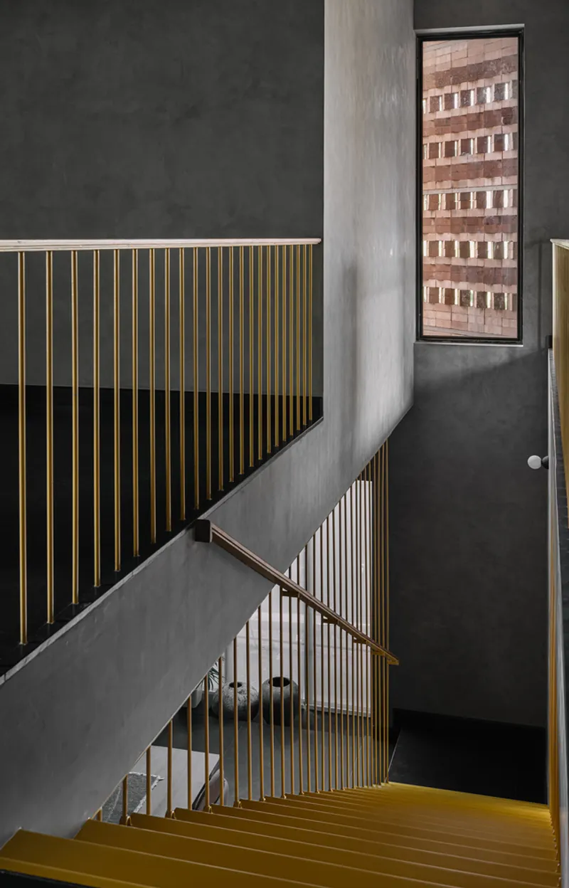 Top-down view of the stairwell showing brass treads, vertical railings, and a tall narrow window framing brick beyond