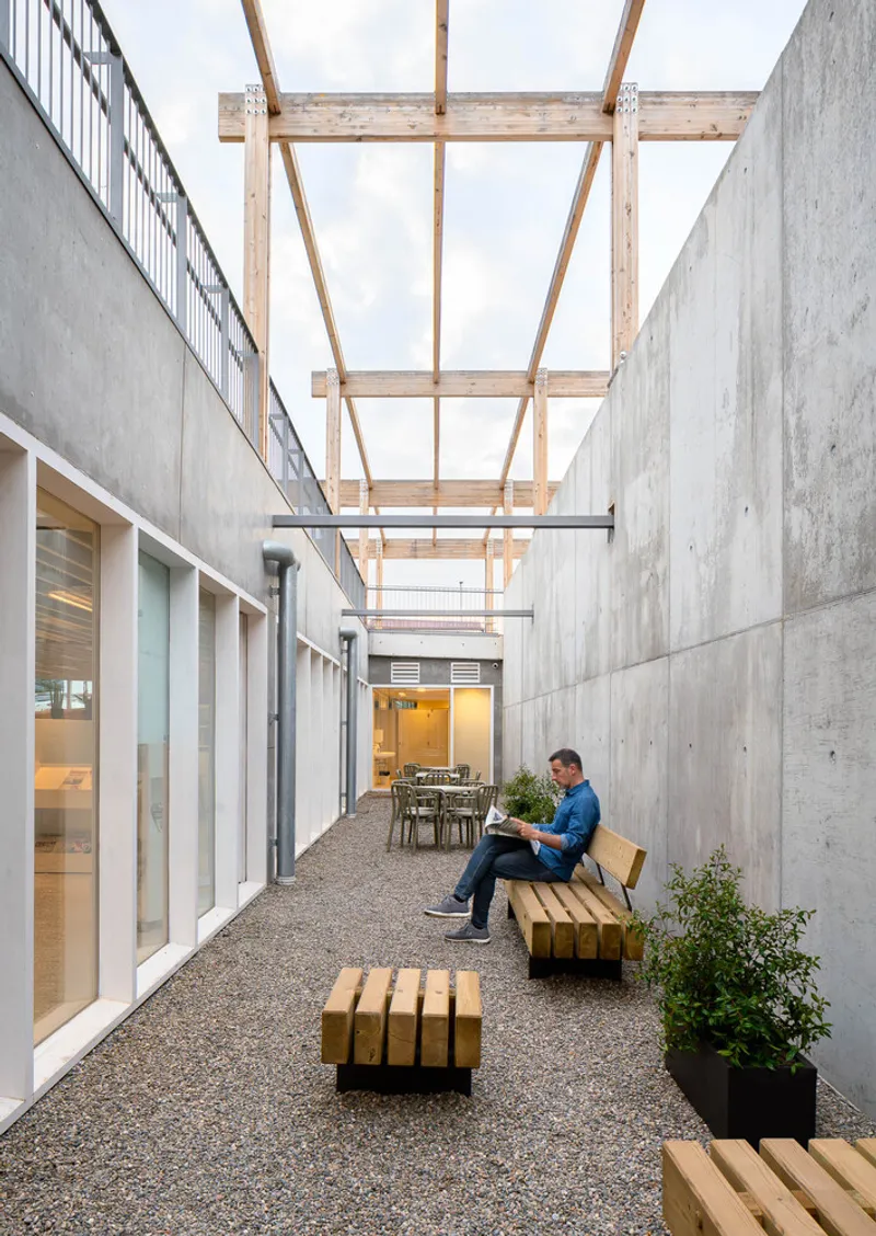 Gravel courtyard flanked by concrete walls with a timber pergola overhead and someone seated on a bench