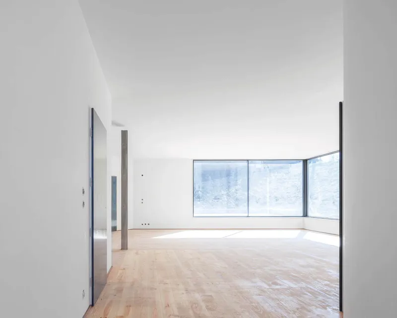 Empty interior room with oak parquet flooring and corner glazing framing views of the concrete retaining wall beyond
