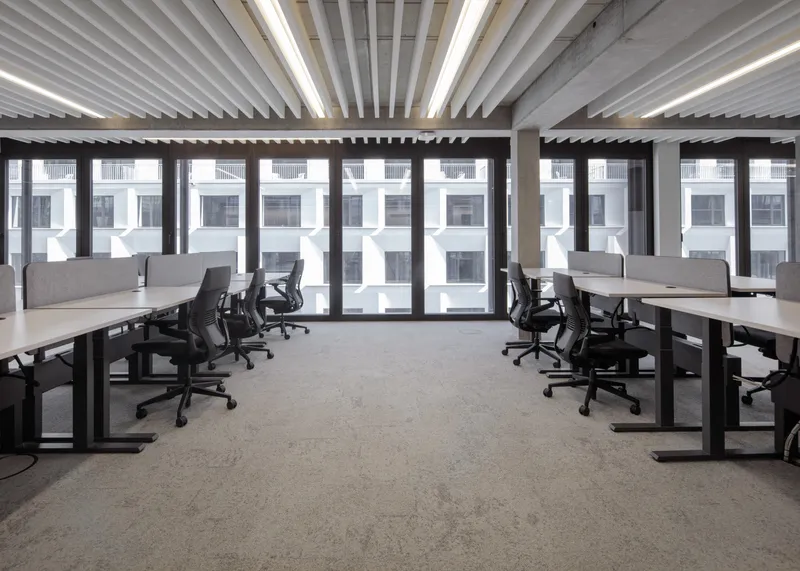 Workspace with rows of desks facing full-height windows under linear ceiling baffles and integrated lighting