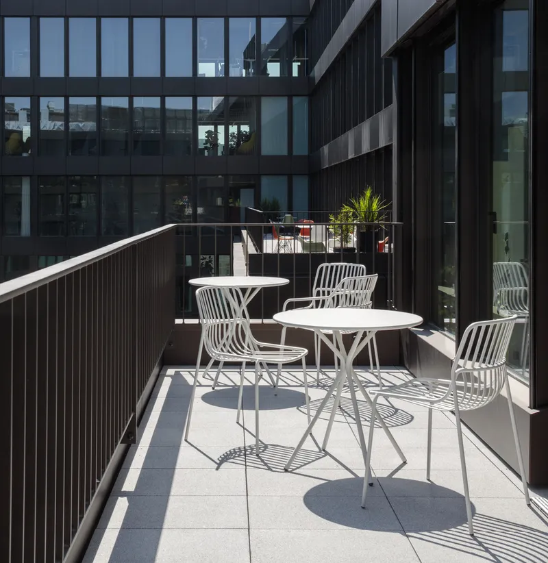 Terrace with white metal tables and chairs in bright sunlight overlooking the dark facade courtyard