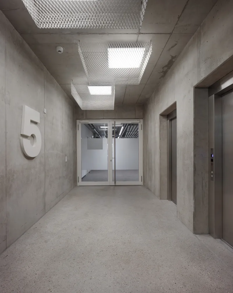 Concrete elevator lobby with mesh-wrapped ceiling light fixtures and glass doors at the corridor end