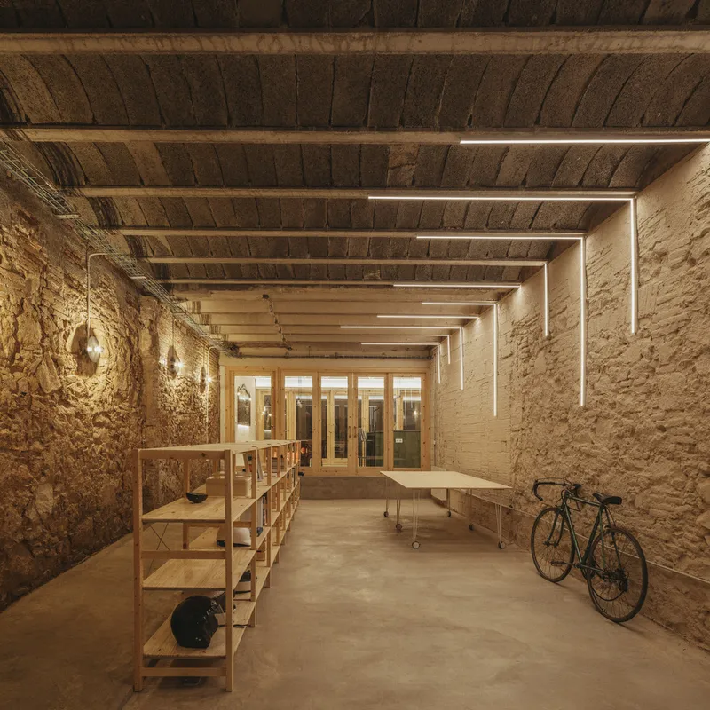 Long workspace with exposed stone walls, timber ceiling beams, linear lighting, and a bicycle