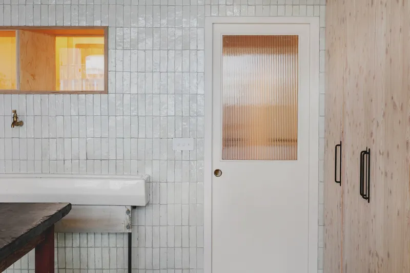 White tiled wall with a door featuring ribbed glass panel beside timber cabinetry with black handles
