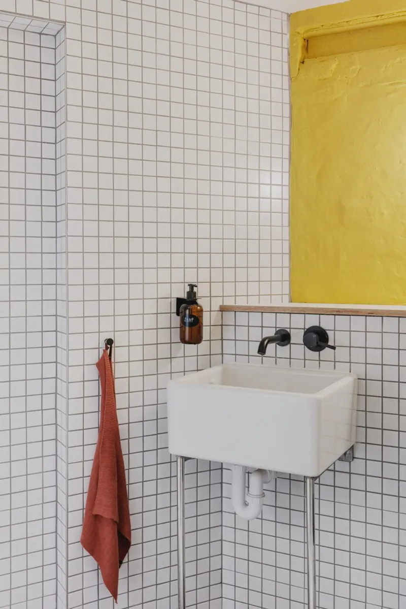 Wall-mounted sink with black faucet beneath a yellow niche on white square tile with an orange towel